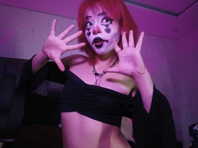 ViolettClay with red hair, white face paint, and black makeup, hands up, black off-shoulder top, exposing her tits, purple light, and a necklace in a dimly lit room.