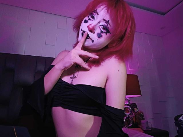ViolettClay, avec ses cheveux roux, son visage peint en blanc, son nez noir et son maquillage, touche ses lèvres avec un doigt, portant un haut noir à une épaule, exposant ses seins..