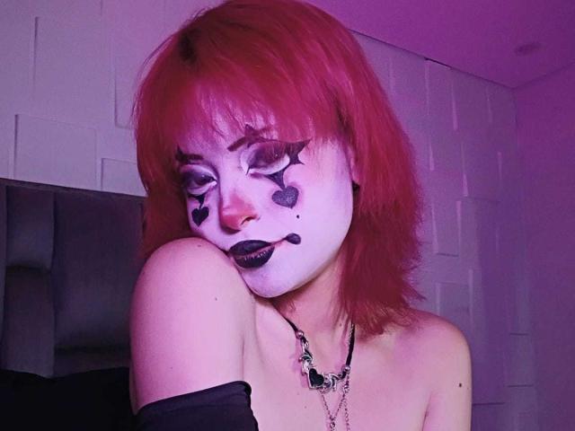 ViolettClay avec des cheveux roses, du maquillage blanc sur le visage, un cœur noir dessiné, du rouge à lèvres foncé, un haut noir à une épaule, un collier, la menton reposant sur l'épaule, une pièce éclairée en violet..