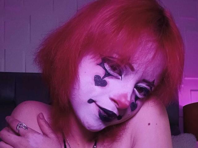 ViolettClay with red hair, white face paint, black heart and lipstick markings, and a ring, leaning on black leather with a purple glow, looking slightly tired.