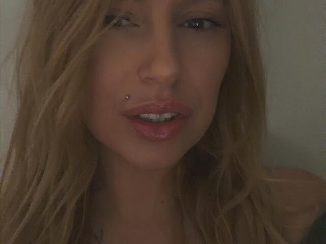 XimenaBella has a pierced nose with a small silver stud, slightly parted pink lips, and blonde wavy hair covering part of her face. Dark background.