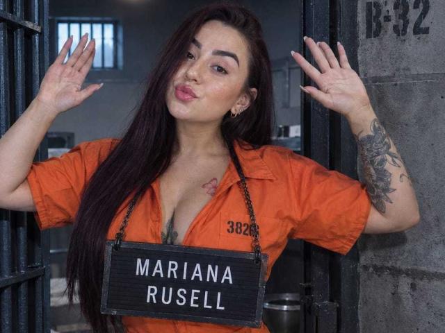 MarianaRusell with long dark hair, pouty lips, orange prison shirt, tattooed arm, holding sign with name, hands up, looking at camera, standing in open prison door.
