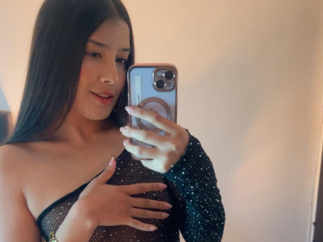 EmmaLombardi taking a selfie in a mirror, wearing a black, see-through mesh dress with sparkly sleeves, covering her tits with one hand, long brown hair, subtle smile.