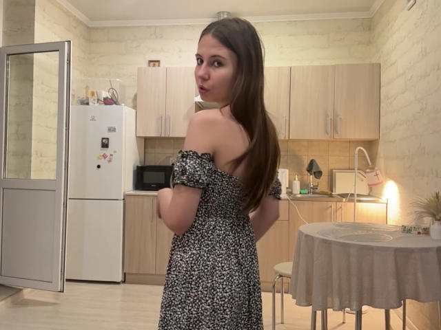 GeorgeCorpeno with long brown hair, off-shoulder black and white floral dress, looking back, showing bare shoulders and upper back, in a kitchen with a round table.