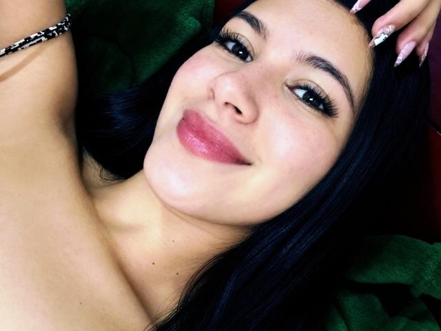 AmilyPreston has smooth skin, dark black hair, and is smiling with pink lips. She has long eyelashes, a black bracelet on her right arm, and is lying down.