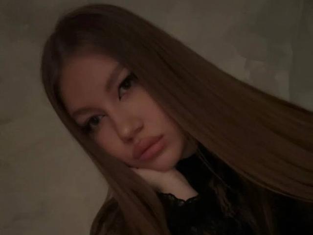 DaniellDuffey with long brown hair, pouty lips, and a serious look, wearing a black lace top, head resting on hand, in a dimly lit setting.