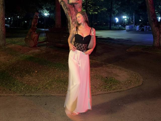 JoelleCalixte with long blonde hair, black strapless top, and white satin pants, standing barefoot in a park at night, holding her hands near her crotch.