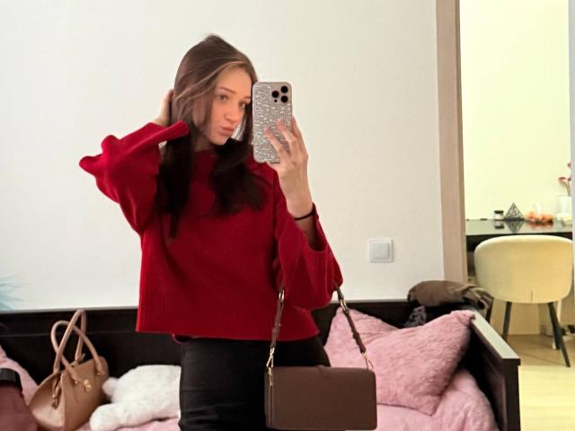 JoelleCalixte with brown hair, red sweater, black pants, holding phone, brown bag, pink pillow, white wall, mirror selfie, hand in hair, looking at phone, bedroom setting.