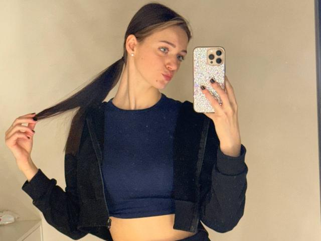 JoelleCalixte, with pigtails, wearing a black jacket and navy crop top, holding her hair, taking a mirror selfie with an iPhone, showing her tits and nose ring.