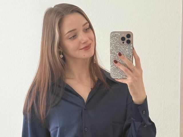 JoelleCalixte with long brown hair, wearing a dark navy shirt, holding a glittery phone, smiling softly, showing a small mole on her right cheek, and red nail polish.