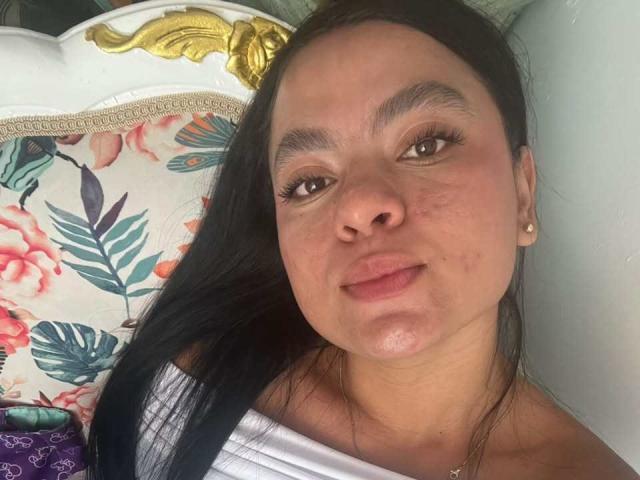 MelanyPorto's face, slightly freckled with dark eyebrows, long black hair, brown eyes, wearing a white off-shoulder top, small pearl earrings, lying on a floral-patterned pillow.
