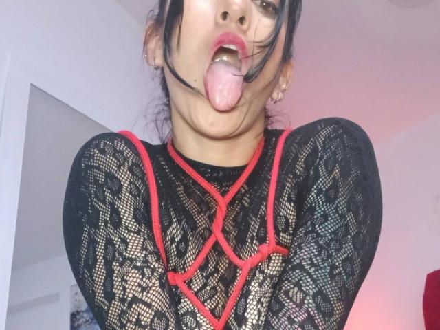 ZarayFeet with tongue out, wearing black lace top with red rope patterns, has dark hair, pale skin, and small earrings. Her mouth is slightly open, tongue sticking out.