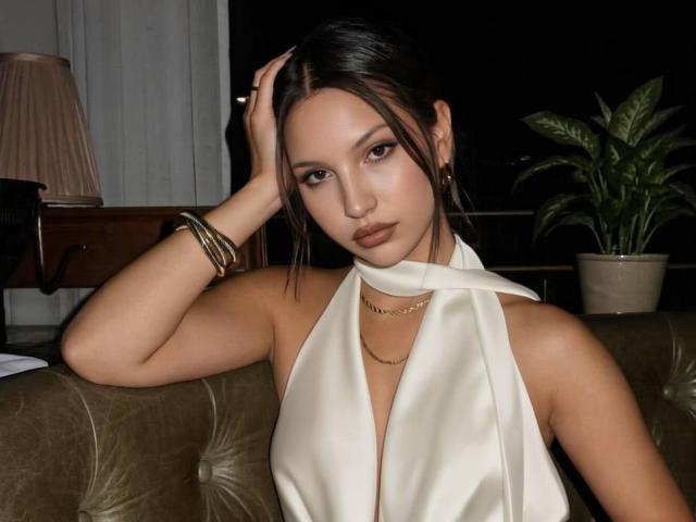 TiarraKoins a une jolie figure, des yeux marron et des cheveux foncés coiffés en demi-queue. Elle porte un haut blanc à encolure dégagée, des bracelets dorés et un collier, et a une main dans les cheveux..