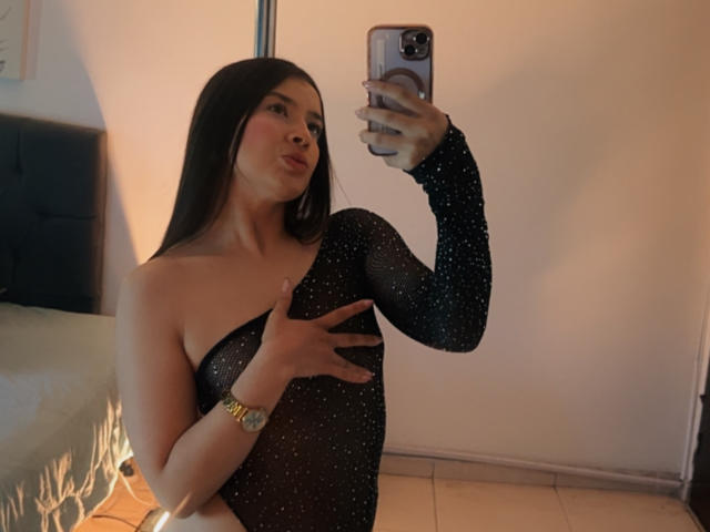 Emma Lombardi mit langem, schwarzem Haar, voller Lippen, trägt ein durchsichtiges schwarzes Kleid mit Pailletten, bedeckt ihre Brüste mit einer Hand und macht ein Selfie vor einem Spiegel mit einer goldenen Uhr..