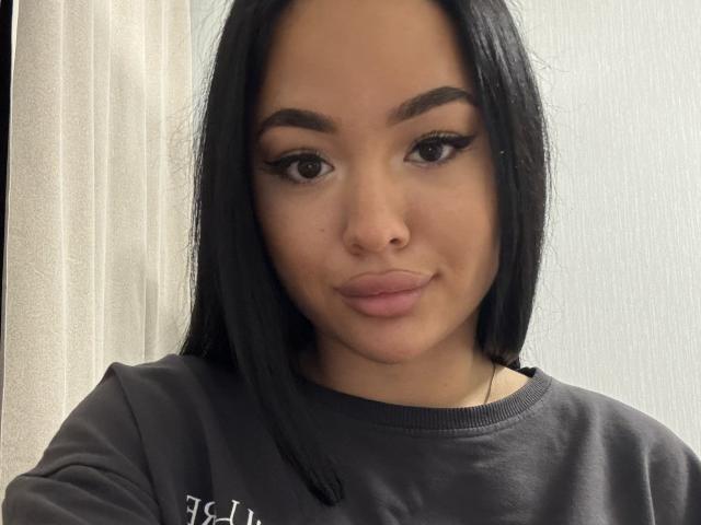 Aletha Bartolomei tem a pele morena, cabelo preto e liso, sobrancelhas grossas, delineador nos olhos e lábios rosados e carnudos. Ela está vestindo um moletom preto e olhando para a câmera com um leve sorriso..