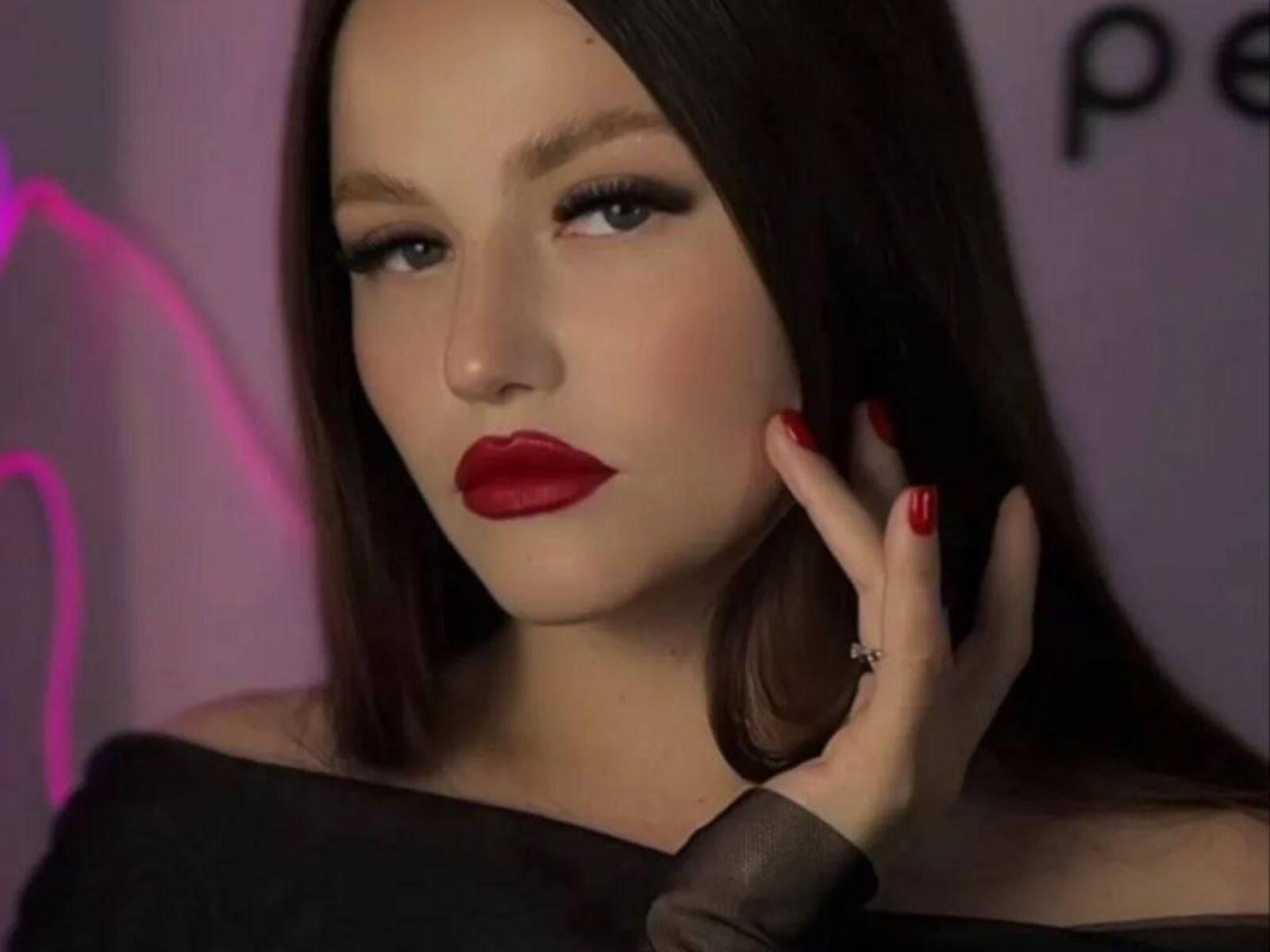 Ethelyn Neher trägt roten Lippenstift, dunkelbraunes Haar und rote Nägel, sie berührt ihr Gesicht mit einem Ring an ihrem Finger und trägt ein schwarzes Oberteil mit einem Schulterauschnitt..