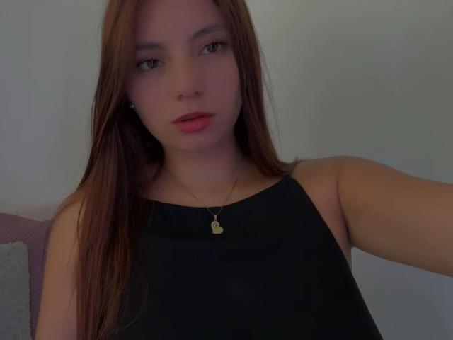LinaFoxyHot has long brown hair, a black sleeveless top, and a gold necklace with a heart pendant. Her pink lips are slightly parted, and she's looking at the camera.