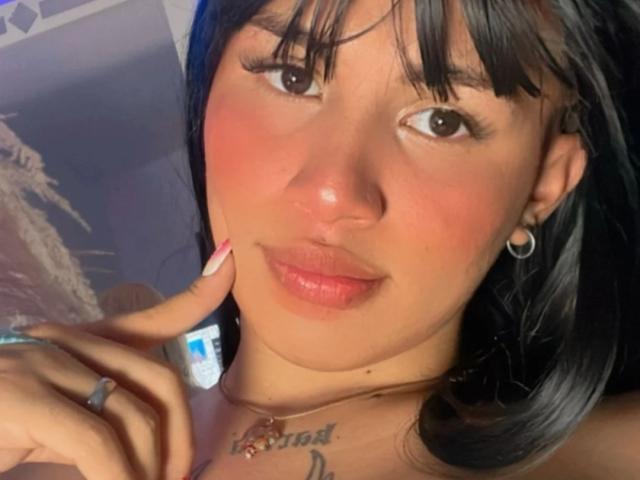 AngelDustdoll, avec ses cheveux noirs, ses lèvres roses et un tatouage sur sa poitrine, touchant sa joue avec ses doigts manucurés de rose, portant des boucles d'oreilles cerceaux et une bague. Elle a un sourire adorable..