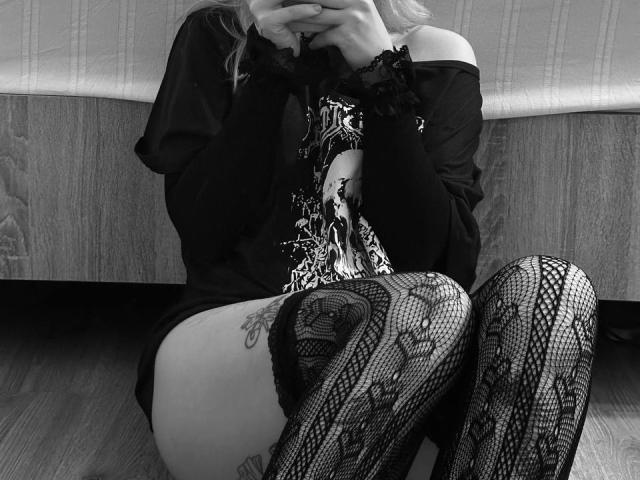 AyumiBabe sitting on floor, wearing off-shoulder black top, lace-patterned thigh-highs, and holding phone to mouth, showing tattoo on right thigh, in black-and-white photo.