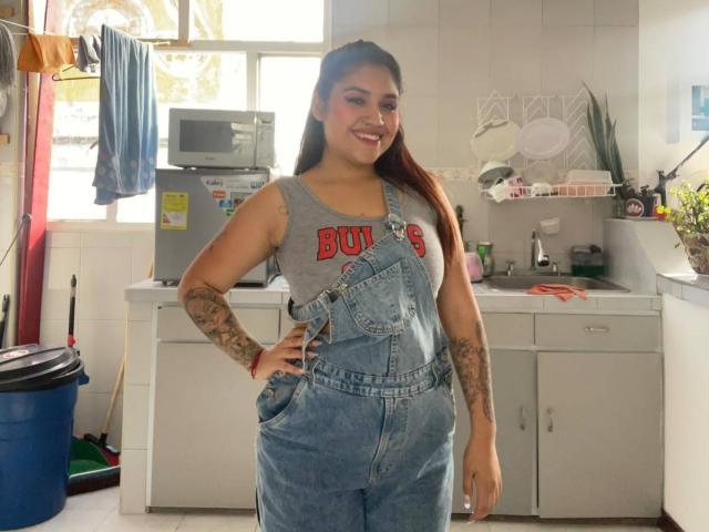 ValmontAlessia with long brown hair, smiling, tattooed arms, wearing denim overalls and a gray "BUNS" tank top, standing in a sunlit kitchen. She's got a cute ass.