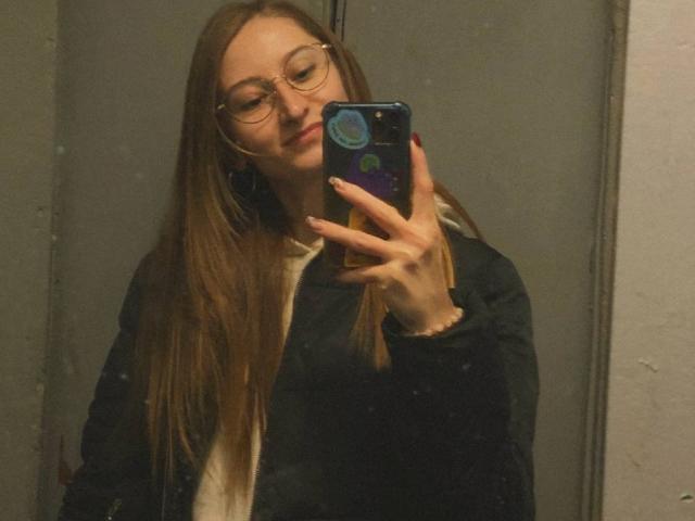 GwynBartell, met lang bruin haar, een bril en een lichte glimlach, maakt een selfie met een telefoon in een donkere kamer, gekleed in een donkere jas over een wit shirt..