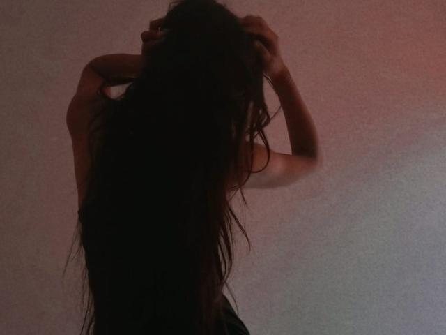 Silueta de GisellRusso con cabello largo y ondulado, de color oscuro, con los brazos levantados, las manos en el cabello, de pie frente a una pared texturizada con una iluminación suave, en un ambiente melancólico y tenuemente iluminado..