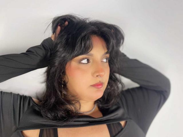 TamaraRousse with black hair, hands in her hair, wearing a black top, looking to the side, pink lips, light skin, small nose, earrings, soft lighting, slightly messy hair.