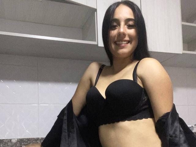 StefanyDelly, avec un soutien-gorge noir, souriante, a une poitrine de taille moyenne, des cheveux noirs, une peau claire, et porte une chemise noire ouverte dans une cuisine. Elle montre son joli visage..