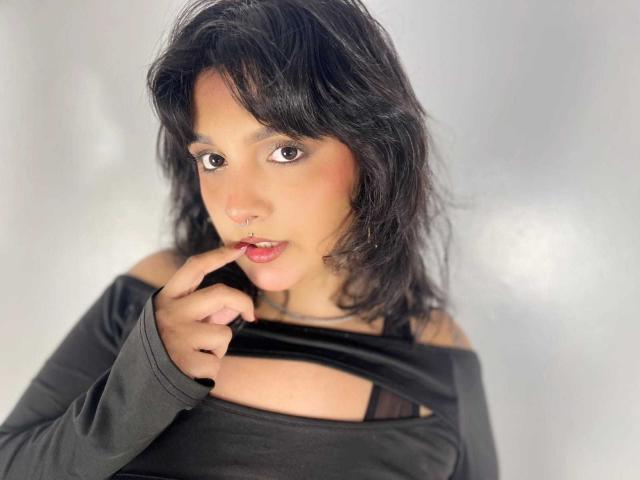 TamaraRousse with black wavy hair, brown eyes, and a nose ring, wearing a black off-shoulder top, biting her finger, looking seductive with a slight smile.