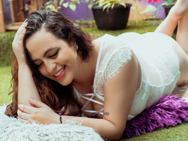 StacyMillerr lying on her stomach, smiling, with wavy brown hair, white lace dress, showing her tits, on a purple pillow, green grass, and a white shaggy rug.
