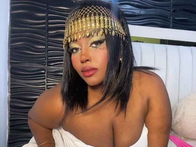 NicolZurich has dark skin, black straight hair, gold chain headpiece, heavy eye makeup, pink lipstick, and large bare boobs, sitting on a white bed with a black textured wall.