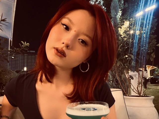 EmaPepper with red hair, hoop earrings, and dark lipstick, holding a drink, leaning slightly forward, looking at camera, night setting, black top, soft lighting.