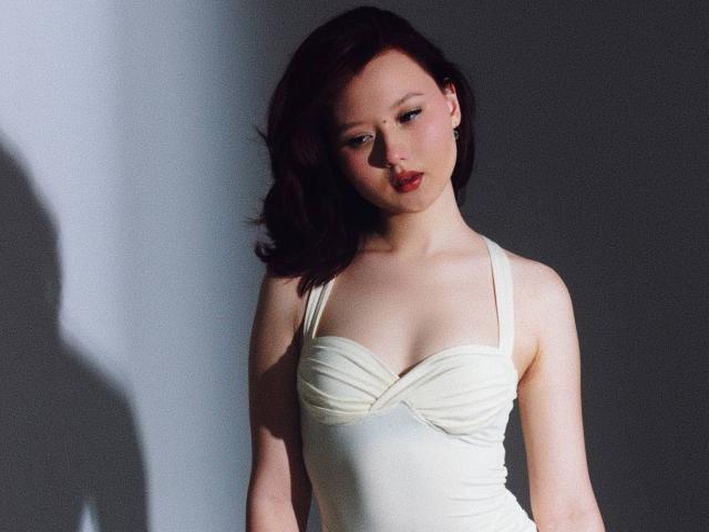 EmaPepper with pale skin, dark red hair, red lipstick, wearing a white, ruched, strapless dress, showing small tits, looking down, shadow on left.