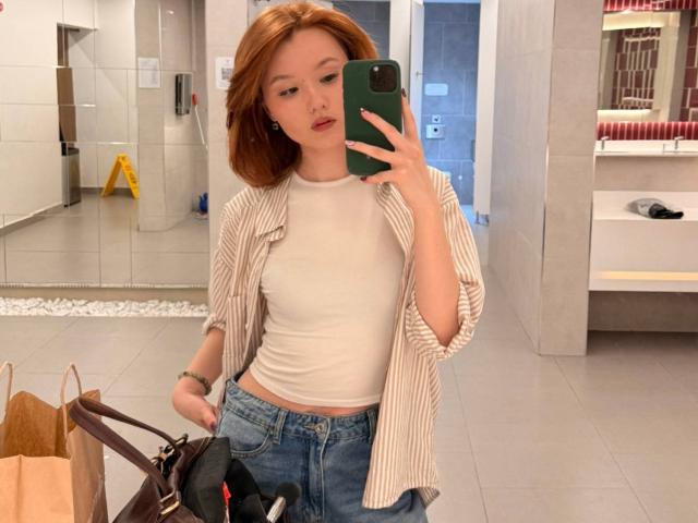 EmaPepper with red hair, white tank top, striped shirt, blue jeans, holding phone, mirror selfie, brown bag, black purse, looking at camera, in a clean, modern bathroom.