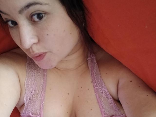 ChiquiAfrodita with brown eyes, pink lace bra, and freckles, lying on an orange pillow, looking at the camera with a slight pout, showing her tits.