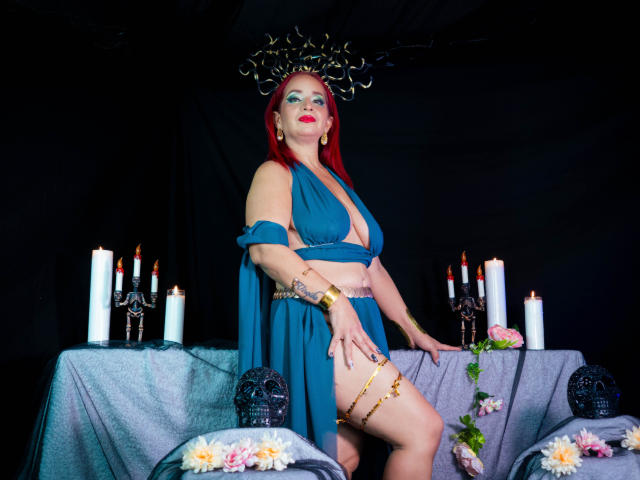 KateMonroe, avec des cheveux roux, un haut de bain bleu à encolure américaine et une jupe assortie, portant une couronne avec des cornes, un bracelet d'or et une jarretière, debout devant des bougies et des fleurs..