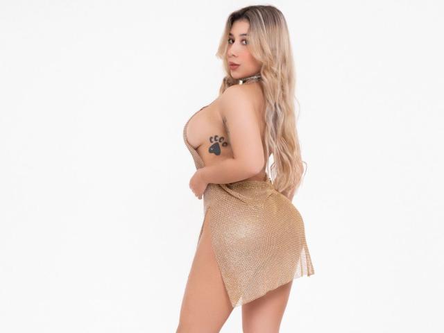 AgostinaNunier in a shiny gold skirt, showing big boobs, a paw tattoo on her side, and a sexy ass, with long blonde hair, looking back over her shoulder.
