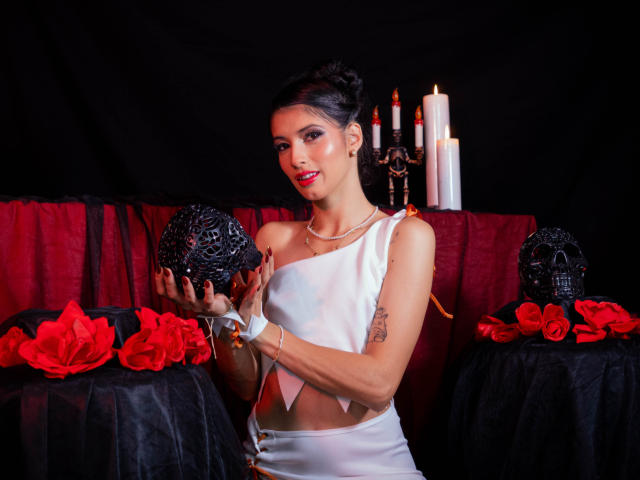 JasminKooper, com cabelo escuro preso em um coque, vestindo um vestido branco de um ombro só, segurando um crânio preto, rosas vermelhas e velas ao fundo, mostrando seus seios..