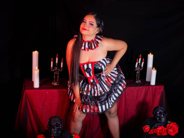 Selena Collins, avec de longs cheveux bruns, du rouge à lèvres, portant une robe rayée comme un zèbre avec des accents rouges, debout devant des bougies et des décorations de crânes, une main sur la hanche, souriant avec assurance..