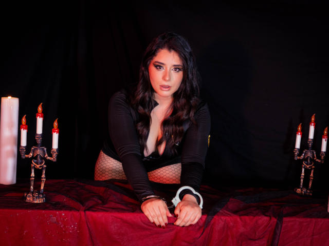 VickyMegan con cabello oscuro y ondulado, vestimenta negra, escote, medias de rejilla, muñequeras sobre la mesa, velas a ambos lados, tela roja, expresión seductora, manos sobre la mesa, llamas rojas de las velas..