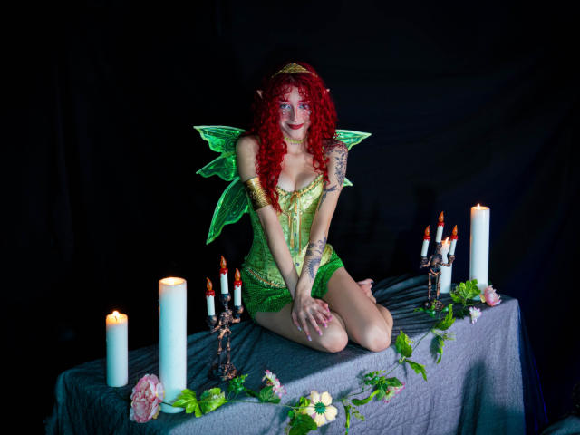 GaiaDoll avec des cheveux roux bouclés, des ailes de fée vertes et un corset en dentelle verte, agenouillée sur une table avec des bougies, des fleurs et un chandelier en métal, dans un décor sombre..