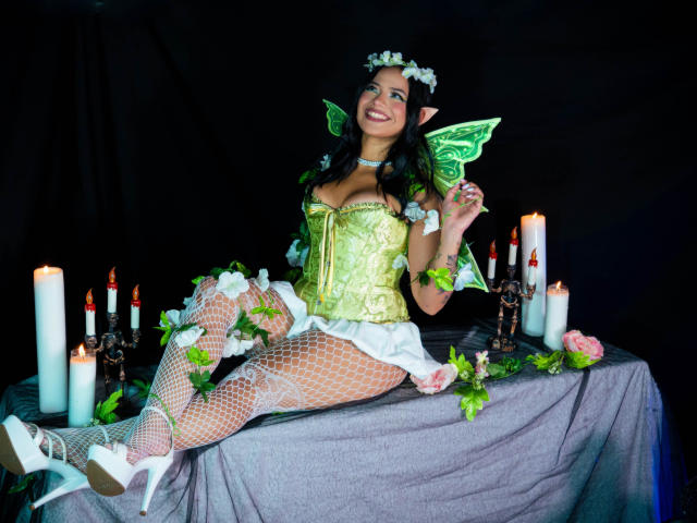 AlanaLuxe, with fairy wings, green corset, white fishnet stockings, and white heels, sits on a table with candles, flowers, and a black background, smiling at the camera.