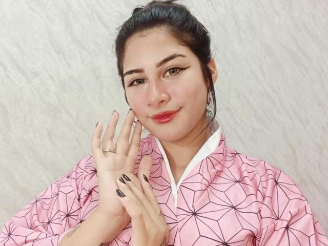 BonnieGray with dark hair in a bun, wearing a pink geometric shirt, has black nail polish, a nose ring, and a ring on her index finger, smiling softly.