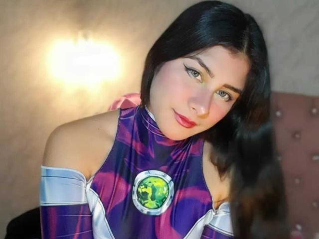 BonnieGray with dark hair, green eyes, and red lipstick, wearing a purple and white tie-dye superhero top, featuring a green gem on her chest, smiling at the camera.