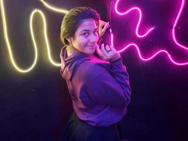 BonnieGray with short brown hair, smiling, wearing a purple hoodie, holding a phone to her ear, neon yellow and pink lights in the background.