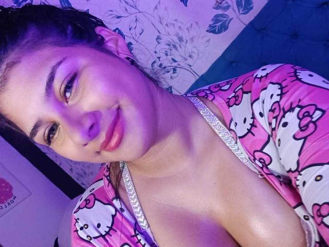 BonnieGray with pink Hello Kitty pajamas, showing cleavage, smiling, purple lighting on face, brown hair in braids, medium skin, cute, small nose, pink lips, visible bra strap.