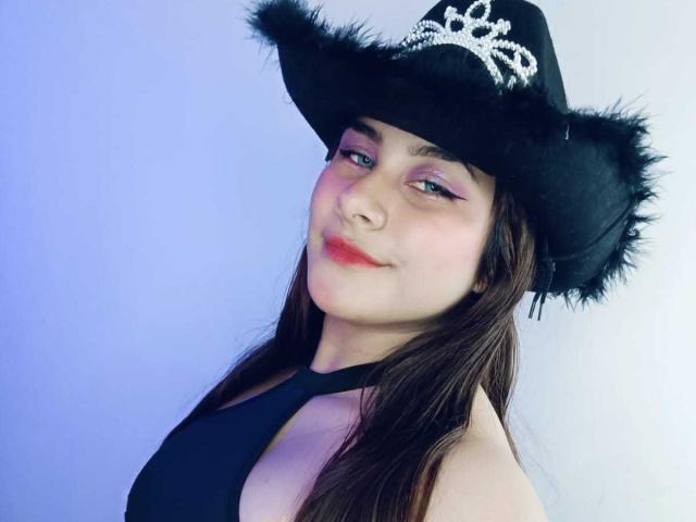BonnieGray with long brown hair, wearing a black cowboy hat with a silver tiara, black halter top, and pink eyeliner, smiling with red lipstick, showing her tits slightly.