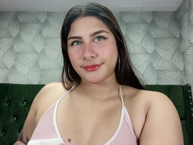 BonnieGray with long dark hair, green eyes, pink glossy lips, wearing a pink and white halter top, showing small nipples, standing against a green tufted headboard.