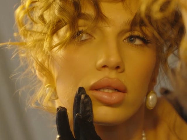 HildeLautner, con cabello rubio rizado, ojos azules, labios rosados brillantes, guantes negros, aretes de perlas, mirando ligeramente hacia un lado, iluminación amarilla, enfoque suave..