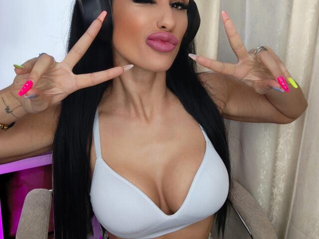 CharlotteAria with big tits in a white bra, long black hair, pink lips, neon nails, and a double peace sign, wearing headphones, in a sexy selfie.